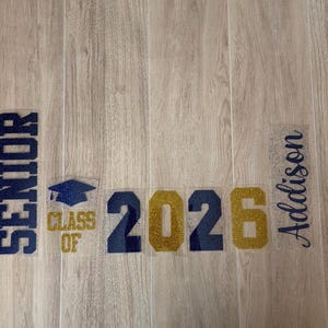 May include: Glittery blue and gold graduation-themed design elements. Includes the word "SENIOR", the text "CLASS OF", a graduation cap, the year "2026", and the name "Addison". The design is on a light wood grain background.