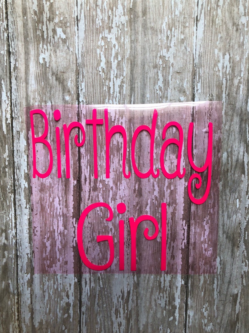 May include: Pink vinyl lettering on a wood background that reads "Birthday Girl".