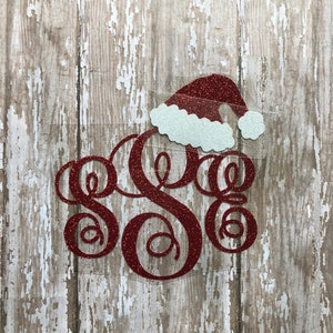 May include: Red glitter monogram with a red and white glitter Santa hat on top. The monogram is on a whitewashed wood background.