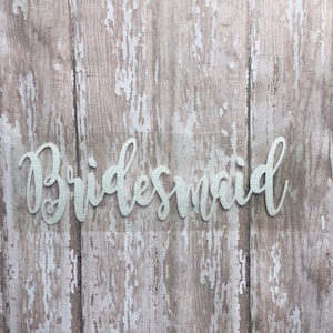 May include: A white glitter "Bridesmaid" sign on a wood background.
