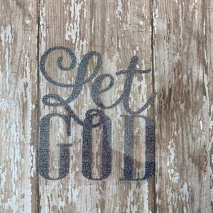 Op de afbeelding: Een zilveren glitterbord met de woorden "Let God" op een rustieke houten achtergrond.