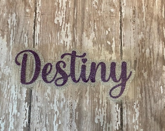 May include: A purple glittery "Destiny" word sticker with a small heart above the "i". The sticker has a silver glitter outline and is set against a weathered wood background.