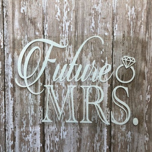 May include: A silver glitter decal featuring the text Future MRS. with a diamond ring icon replacing the letter O. The lettering is presented in a combination of cursive and serif fonts, designed for application onto a smooth surface.