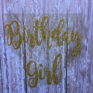 May include: Gold glitter birthday girl sign on a wood background.