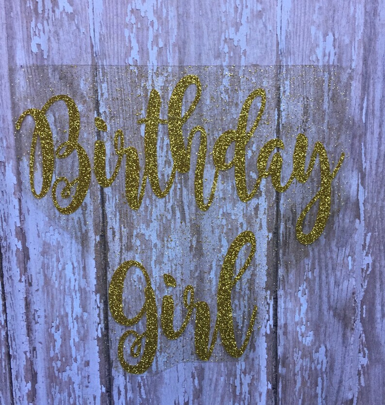 May include: Gold glitter birthday girl sign on a wood background.