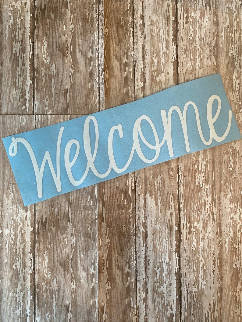 Op de afbeelding: Een lichtblauw welkom bord met witte, cursieve letters. Het bord is op een houten achtergrond.