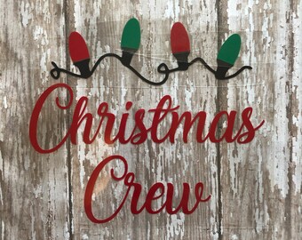 May include: Red and green Christmas lights strung together with the words "Christmas Crew" in red lettering on a wood background.