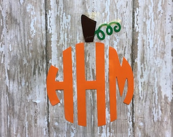 May include: An orange vinyl decal shaped like a pumpkin featuring the monogram letters HHM in a stylized font. The design includes a dark brown stem and a green curly vine detail at the top, all applied to a clear adhesive backing sheet.