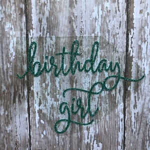 May include: Green glitter birthday girl sign on a wood background.