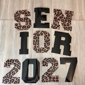 May include: A set of iron-on heat transfer decals spelling out SENIOR 2027. The letters and numbers alternate between a black glitter finish and a tan and brown leopard print pattern, arranged in three rows on a clear backing sheet.
