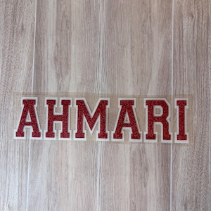 May include: A custom iron-on transfer decal featuring the name AHMARI in a bold, collegiate-style block font. The letters are rendered in a vibrant red glitter finish with a thin white outline, all set against a clear, transparent adhesive backing.