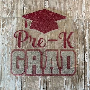 May include: A glittery red graduation cap and the words "Pre-K GRAD" in a silver and red design. The image is on a weathered, light-colored wooden background, perfect for a graduation celebration.