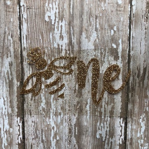 May include: Gold glitter "Bee" and "Me" cutout letters on a weathered wood background.