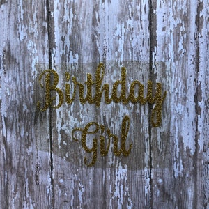 May include: Gold glitter letters spelling "Birthday Girl" on a weathered wood background.