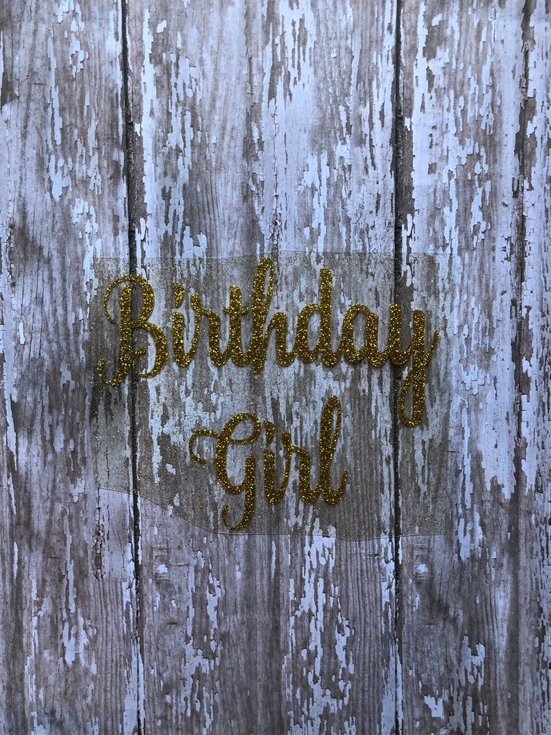 May include: Gold glitter letters spelling "Birthday Girl" on a weathered wood background.