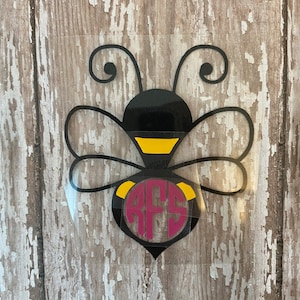May include: A black and yellow bee with a pink monogram in the center. The bee has a black body with yellow stripes and a pair of black antennae.