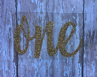 May include: A gold glitter script word cutout that spells out the word one. The letters are connected in a cursive font style and feature a textured, sparkling surface that reflects light across the entire surface of the decorative piece.