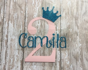 May include: Pink number two with a teal glitter crown and the name "Camila" in teal glitter.