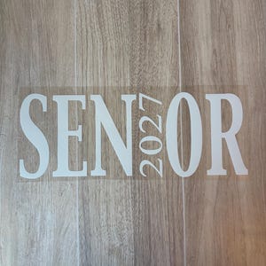 May include: A white vinyl decal featuring the word SENIOR in a bold, serif font. The numbers 2027 are stacked vertically in place of the letter I, creating a cohesive design. The decal is presented on a clear transfer sheet for application.