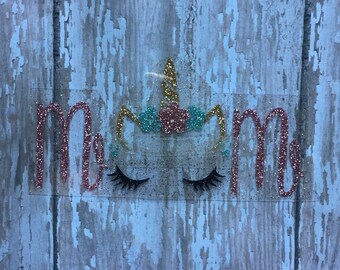 May include: A glittery "Mom" design with a unicorn face, including a gold horn, teal and pink accents, and black eyelashes. The design is on a clear background.