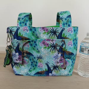 May include: A teal tote bag with a peacock and floral pattern. The bag has two handles and two front pockets. A pair of eyeglasses is in one pocket, and a set of keys is attached to the side. A water bottle is next to the bag.