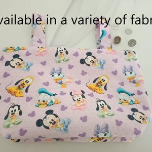 May include: A pink fabric tote bag featuring a variety of Disney characters printed on it. The characters include Mickey Mouse, Minnie Mouse, Donald Duck, Goofy, Pluto, and Daisy Duck. The bag has a pink background with white polka dots and white hearts. The text "Available in a variety of fabrics" is printed at the top of the bag.
