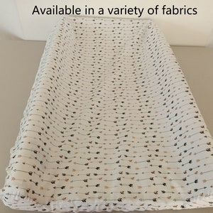 Puede incluir: Una funda de cambiador blanca con un patrón de flechas marrones y negras. "Disponible en una variedad de telas" está escrito en la parte superior de la imagen.