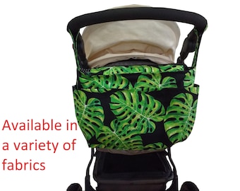 May include: A stroller organizer with a black and green tropical leaf pattern. The organizer has multiple pockets and a black handle. The text "Available in a variety of fabrics" is visible in the image.