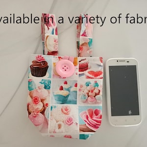 May include: A small tote bag with a cupcake pattern, featuring a large pink button closure and matching handles. A white smartphone is placed next to the bag. Text on the image reads "Available in a variety of fabrics."