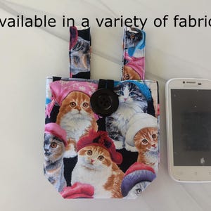 May include: A small tote bag with a black button closure and two handles. The bag is decorated with a colorful cat print, and the text "Available in a variety of fabrics" is visible. A white smartphone is next to the bag.