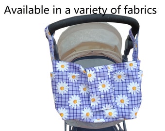 May include: A stroller organizer bag with a daisy floral pattern on a purple and white checkered background. The bag has multiple pockets and is attached to the stroller handle. Text on the image reads: Available in a variety of fabrics.