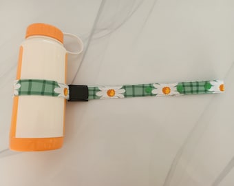 Könnte beinhalten: Eine weiße Wasserflasche mit orangefarbenem Deckel und einem grün-weißen Gurtband mit Gänseblümchenmuster und einer schwarzen Schnalle.