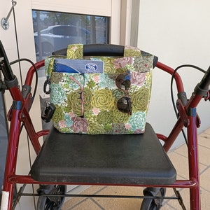 May include: A green and brown floral patterned tote bag with multiple pockets sits on the seat of a red walker. The bag has a zipped pocket and a key clip. Sunglasses are visible in the pockets.