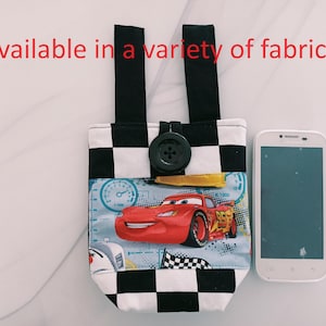 Puede incluir: Una funda para teléfono de tela a cuadros en blanco y negro con un diseño de coche de carreras rojo y un botón negro. La funda tiene dos correas negras para llevar. "Disponible en una variedad de telas" está escrito en rojo en la parte superior de la imagen.