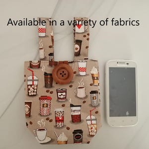 May include: A small, beige fabric tote bag with a brown button closure. The bag is decorated with a coffee cup pattern and the text "Available in a variety of fabrics".