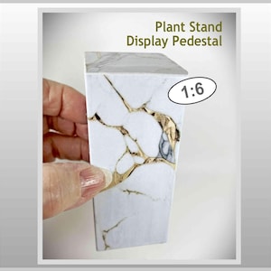 Puede incluir: Un pedestal de exhibición blanco y dorado con el texto "Plant Stand Display Pedestal" y "1:6". El pedestal tiene un patrón similar al mármol con vetas doradas. Está diseñado para exhibir plantas u otros objetos pequeños.