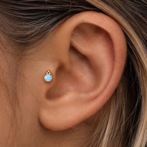 Op de afbeelding: Een gouden tragus-oorbel met een kleine, ronde, lichtblauwe steen in een zetting, bekroond met drie kleine gouden bolletjes in een driehoekige opstelling. Het sieraad bevindt zich in het tragus-gebied van het oor en heeft een metalen afwerking.