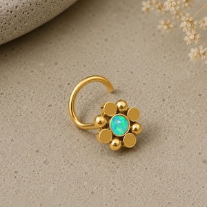 Può includere: Un anello da naso color oro con un design a forma di fiore. Il centro del fiore è una pietra di opale rotonda blu-verde iridescente, circondata da sei piccole sfere lisce color oro disposte in un motivo circolare su un filo curvo.