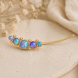 May include: A gold-toned ear climber earring featuring five round, iridescent blue and purple opal-like stones of varying sizes, arranged in a curved line with small gold spheres between each stone and a long, slender gold wire backing.