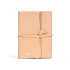 Puede incluir: Un cuaderno de cuero beige claro con una correa de cuero anudada y la palabra "INK" grabada en la parte delantera.