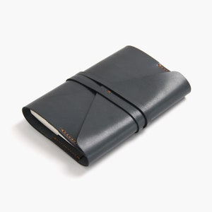 Puede incluir: Un cuaderno de cuero gris oscuro con una correa y costuras de cuero marrón. El cuaderno está cerrado y se encuentra sobre una superficie blanca.