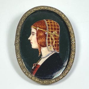 Peut inclure: Broche ovale avec un cadre en métal doré. Le cadre présente un motif floral détaillé. À l'intérieur du cadre, un portrait peint d'une femme aux cheveux roux portant un couvre-chef blanc et rouge. L'arrière-plan est vert foncé.