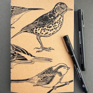 May include: A brown notebook with black bird illustrations and two black pens. The notebook features detailed drawings of various birds. The pens are black with white text. The notebook is open, displaying the bird drawings.