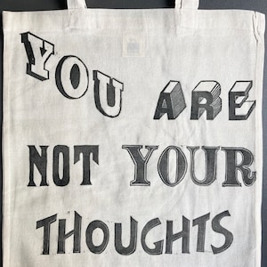 May include: A white canvas tote bag with black text that reads "You are not your thoughts". The text is in a bold, blocky font.