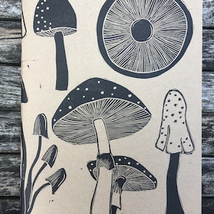 Puede incluir: Una impresión de linóleo en blanco y negro de setas sobre un fondo de papel marrón. La impresión presenta una variedad de setas, incluyendo una seta grande con un sombrero ancho, una seta más pequeña con un sombrero puntiagudo y un grupo de setas pequeñas.