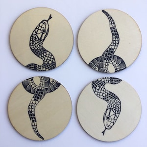 Può includere: Set di quattro sottobicchieri in legno con illustrazioni di serpenti neri. Ogni sottobicchiere presenta una posa diversa del serpente, con squame e motivi dettagliati. I sottobicchieri hanno uno sfondo in legno naturale.