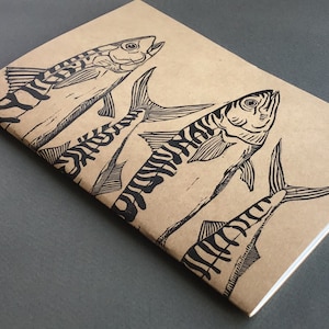 May include: A brown notebook with a black and white illustration of three fish. The fish are drawn in a stylized, almost cartoon-like way, with large eyes and fins.