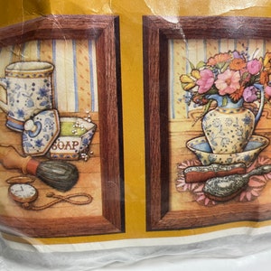 May include: Two framed cross-stitch patterns, one depicting a blue and white mug, soap dish, and shaving brush, and the other a blue and white vase with flowers and a hairbrush. The text on the soap dish reads "SOAP".