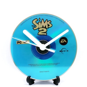 May include: A blue and white clock made from a The Sims 2 PC game disc. The clock face features the game's logo and the EA and Maxis logos. The clock hands are white.