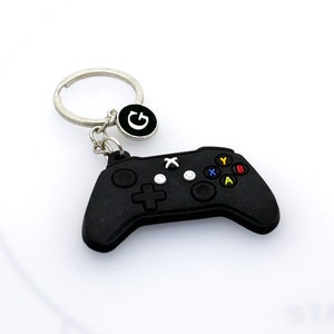 May include: Black video game controller keychain with a silver keyring and a silver charm with the letter "G" in white.